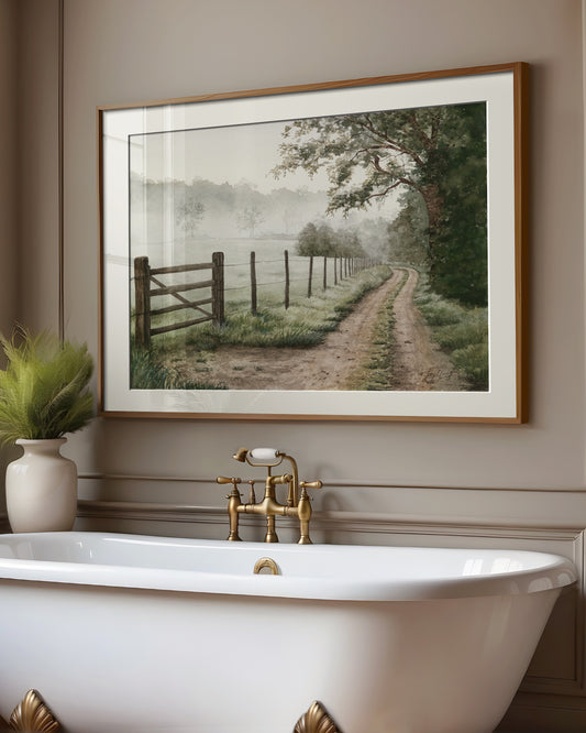 Framed landscape print of an Australian country side where there is a gate leading into a misty paddock and a path leading into the distance above a freestanding bathtub with gold fixtures.