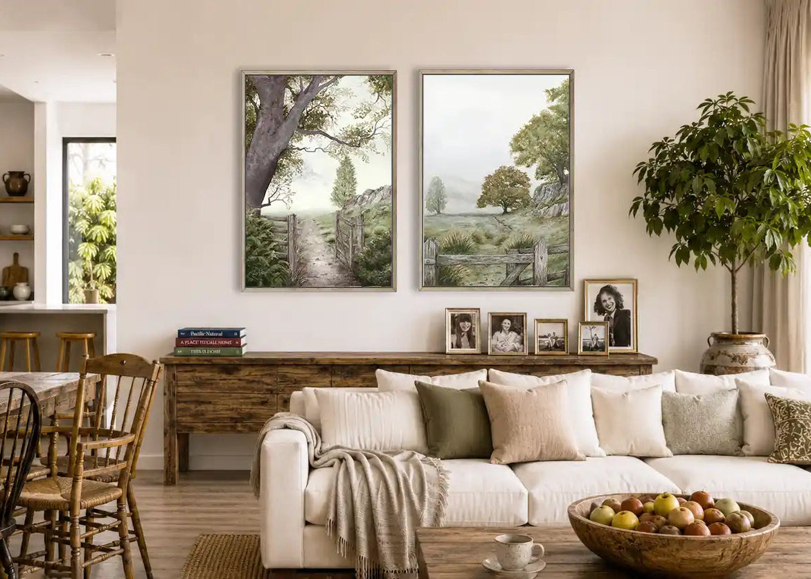 A country Australian styled living room interior featuring two framed landscape art prints hanging on a cream wall above a wooden console table, with a white sofa, dining area, and a potted plant in the background.